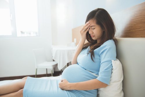 Dor de cabeça na gravidez o que fazer Mulher grávida na cama sentindo forte dor de cabeça.