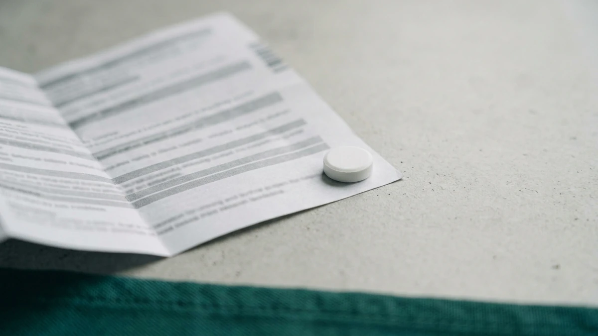 Bula de papel dobrada e aberta sobre uma superfície clara, com o texto impresso fora de foco e ilegível, e um comprimido branco redondo apoiado sobre a borda do papel