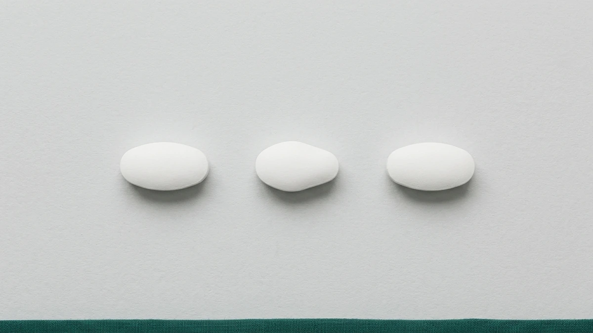 Três comprimidos brancos de formatos ligeiramente diferentes alinhados lado a lado sobre superfície cinza clara, igualmente espaçados e igualmente iluminados, com faixa verde-escura na borda inferior